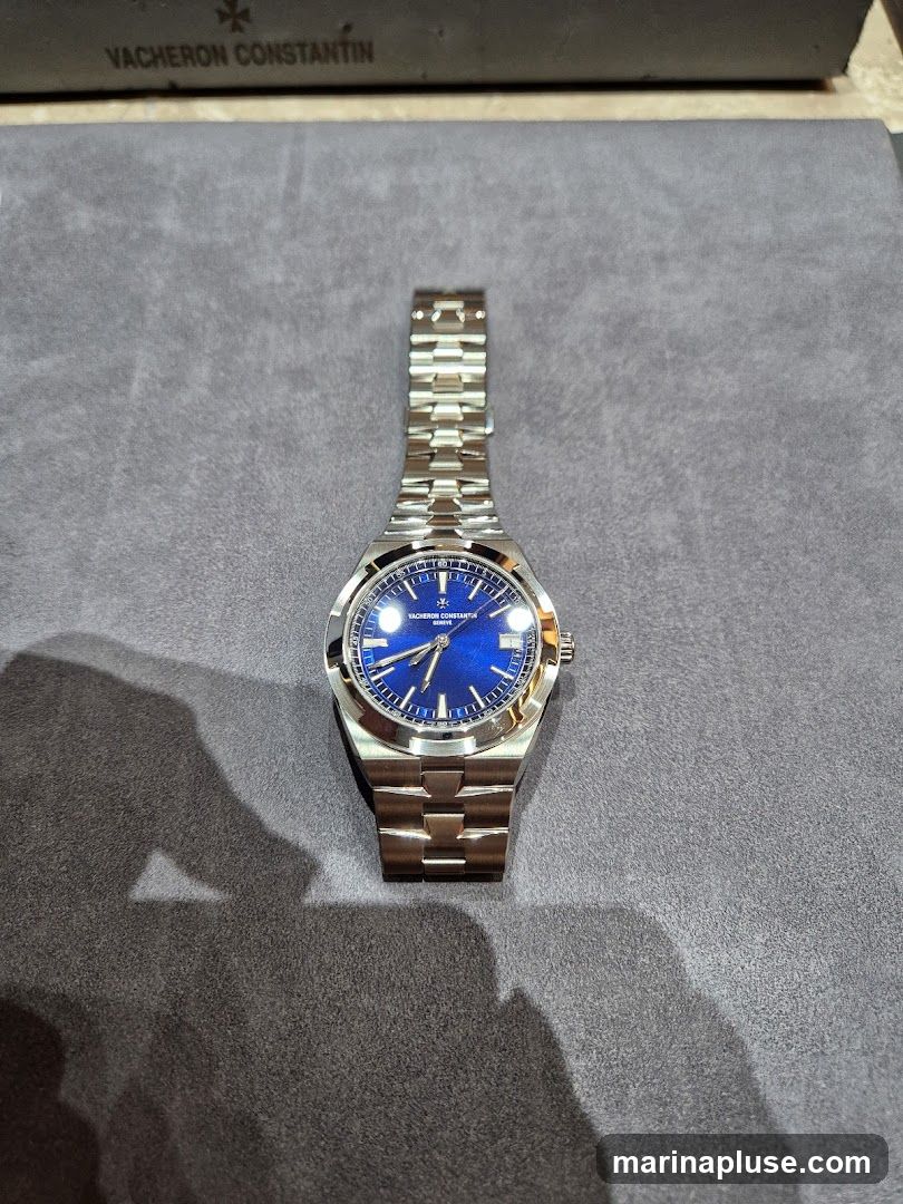 A close-up shot of a Vacheron Constantin watch with a blue dial and a stainless steel bracelet. The watch is placed on a textured gray surface. The Vacheron Constantin logo and name are visible on the watch face and on a dark gray box in the background.