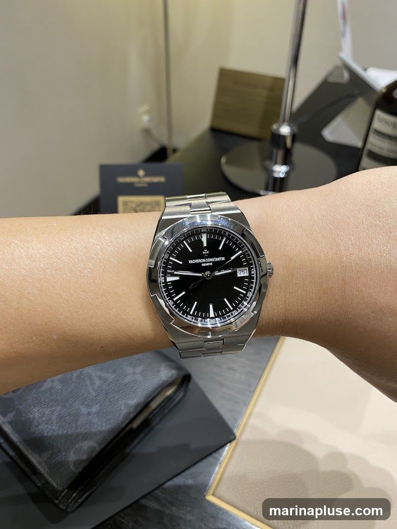 A close-up shot of a Vacheron Constantin watch on a person's wrist. The watch has a black dial with silver hour markers and hands, and a date window at the 3 o'clock position. The watch is made of stainless steel with a metal bracelet. In the background, there is a dark surface with a wallet and a small card with a QR code and the Vacheron Constantin logo.