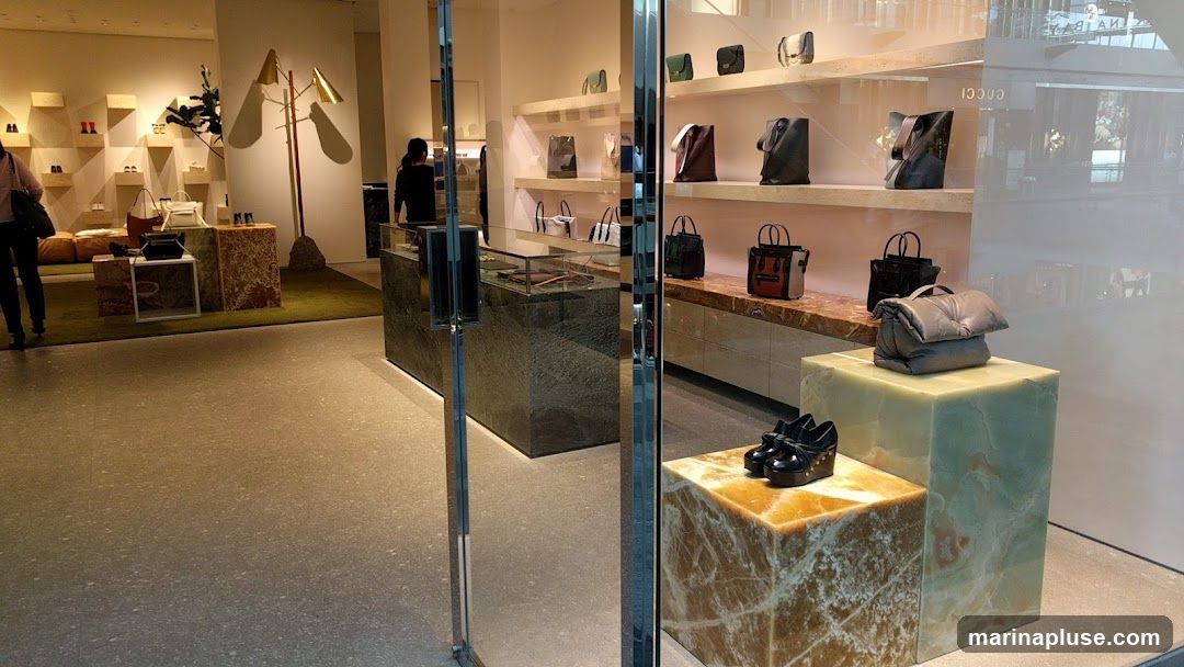 A well-lit interior of a luxury retail store, showcasing a collection of designer handbags and shoes. The display features marble pedestals and shelves, with handbags arranged neatly on shelves and a plush grey handbag prominently displayed on a light green onyx cube. A pair of black wedge heels with gold accents are showcased on a smaller, amber-hued marble cube. In the background, more handbags and shoes are visible, along with a seating area and a modern floor lamp. The store appears to have a clean and minimalist aesthetic.