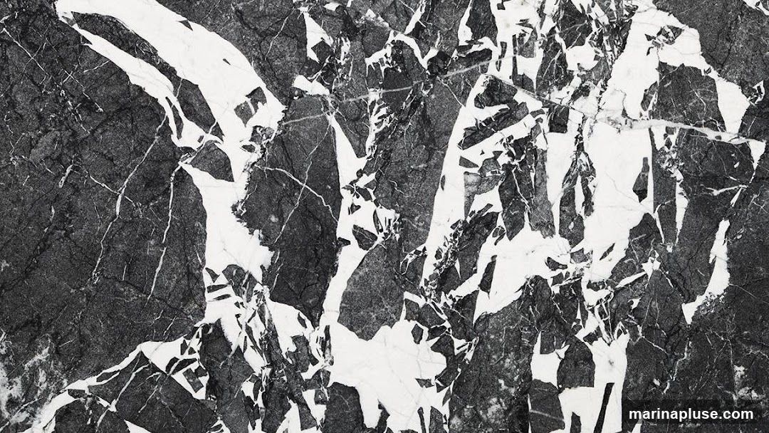 A close-up, abstract view of a black and white marble surface, showcasing its natural veining and crystalline structure. The pattern is characterized by bold, irregular white patches and streaks against a dark, charcoal-grey background, with fine white lines crisscrossing the darker areas.