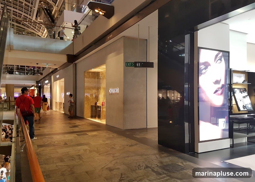 A view of a Celine store with its name displayed prominently on the facade. The store features a large glass window showcasing handbags. To the right, a large digital screen displays a close-up of a woman's face, likely an advertisement. The surrounding area appears to be a shopping mall with other stores and escalators visible in the background.