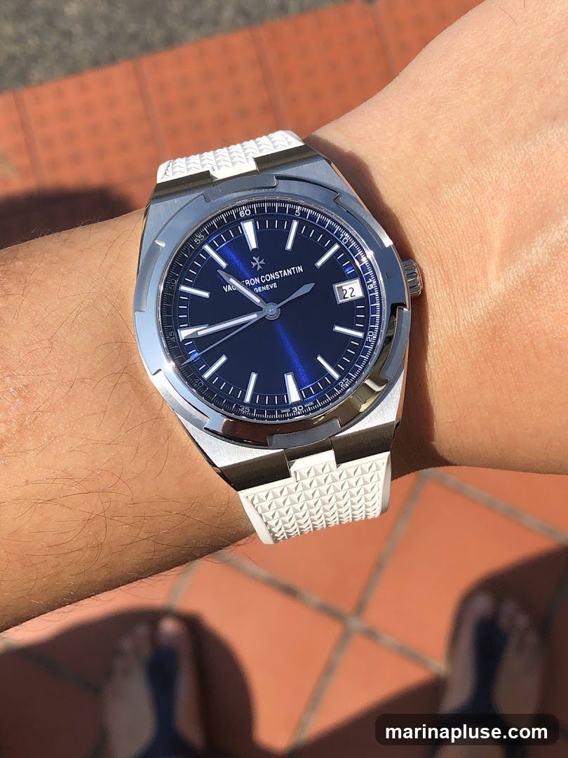 A close-up shot of a Vacheron Constantin watch on a person's wrist. The watch has a stainless steel case and a blue dial with silver hour markers and hands. The date window is visible at the 3 o'clock position. The watch band is white with a textured pattern. The background shows a tiled surface and a person's feet wearing sandals.