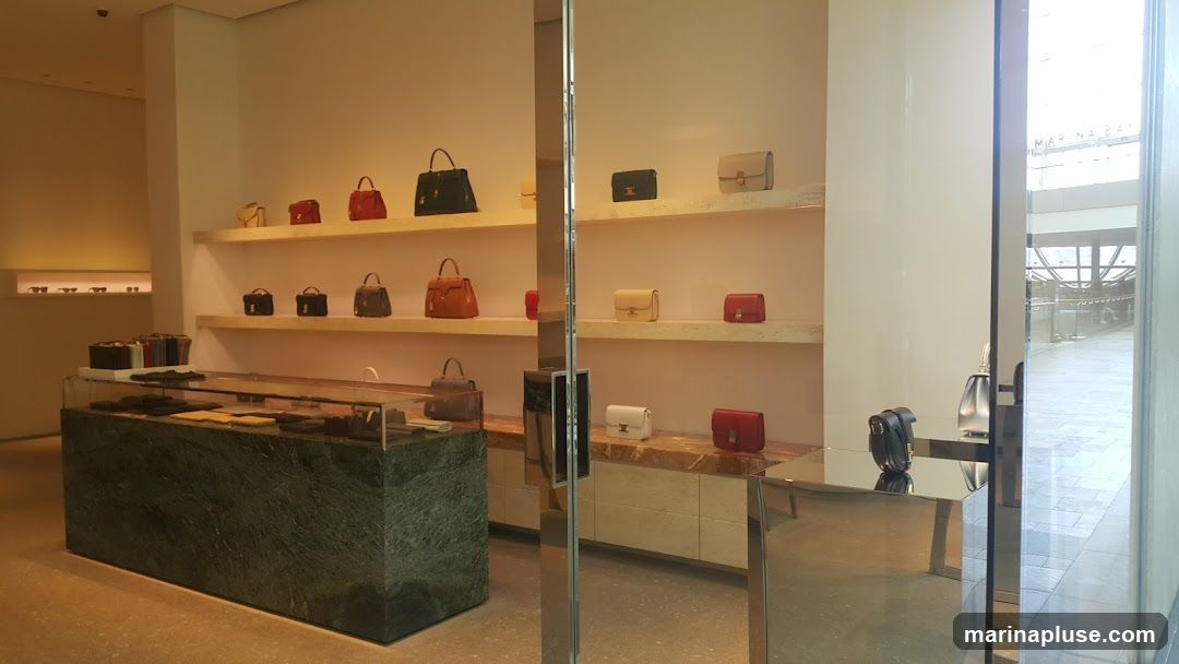 The interior of a luxury store displays various handbags on shelves and in a display case. The store features a modern and minimalist design with light-colored walls and shelves. A large, dark green marble counter is positioned in the foreground, showcasing smaller items. The overall ambiance is elegant and sophisticated, with soft lighting highlighting the merchandise. Reflections from polished surfaces add to the luxurious feel. Through the open doorway, a glimpse of a mall interior with tiled flooring and architectural elements is visible.
