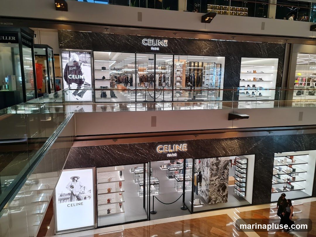 Two Celine storefronts are visible in a shopping mall. The upper storefront displays clothing and accessories, with a large advertisement featuring a person wearing a Celine jacket. The lower storefront showcases handbags and shoes, with a prominent display of the Celine logo and branding. Both stores have a modern and luxurious aesthetic, with marble accents and well-lit displays.