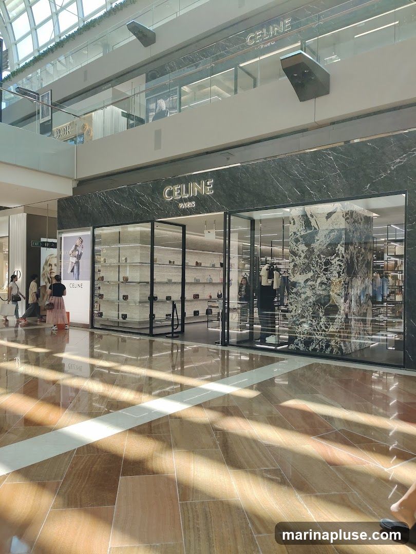 A well-lit, modern storefront for Celine Paris, featuring a marble facade and large glass windows displaying handbags and clothing. The store is located in a multi-level shopping mall with a polished wooden floor reflecting the sunlight.