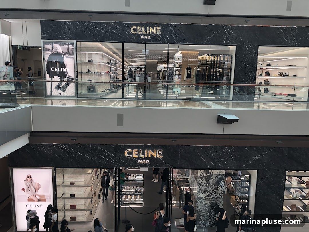 The image shows the exterior of a Celine store located in a mall. The store has a black marble facade with the Celine Paris logo prominently displayed in gold lettering. Large glass windows reveal the interior displays of clothing, handbags, and accessories. There are also large advertisements featuring models wearing Celine products. People are seen entering and browsing the store.