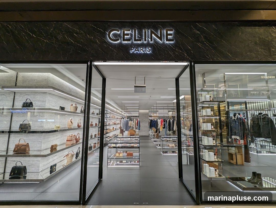 The exterior of a Celine Paris store. The entrance is wide and open, revealing a well-lit interior with displays of handbags, clothing, and accessories. The store's name, "CELINE PARIS," is prominently displayed in illuminated lettering above the entrance. The facade features sleek, modern design with marble accents and large glass windows.
