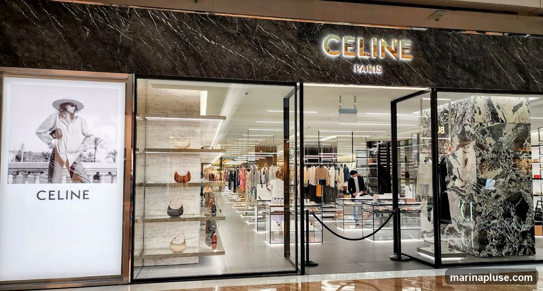 The exterior of a Celine Paris store. The facade features a large illuminated advertisement on the left with the Celine logo and a fashion image. The main storefront has large glass windows and doors, revealing a well-lit interior with clothing racks, shelves displaying handbags, and a marble-accented pillar. The Celine Paris logo is prominently displayed above the entrance.