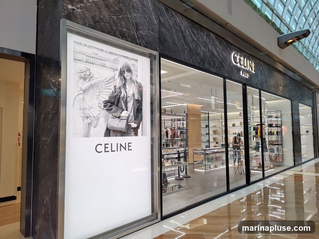 A wide shot of the exterior of a Celine store. The store has a dark marble facade with large glass windows. A large advertisement featuring a woman wearing sunglasses and holding a handbag is displayed on the left side of the storefront, with the word "CELINE" printed below it. The Celine logo is visible above the entrance. Inside the store, racks of clothing and shelves displaying handbags and accessories can be seen.