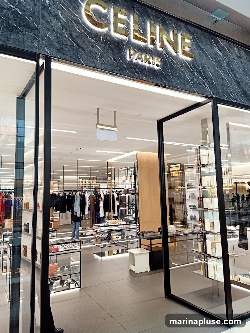 The exterior of a Celine Paris store, featuring a marble facade with illuminated gold lettering spelling out "CELINE PARIS". The entrance is open, revealing a well-lit interior with clothing racks, display shelves, and merchandise.