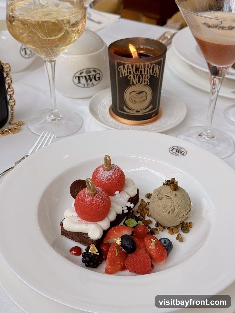 A white plate holds a chocolate dessert topped with red spheres and whipped cream, accompanied by fresh berries and a scoop of ice cream. A lit candle with "MACARON NOIR" branding sits nearby, along with a glass of white wine and a cocktail. A sugar pot with the TWG Tea logo is also visible.
