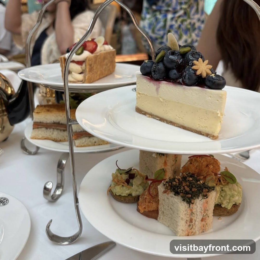 A tiered stand holds a selection of delicate pastries and sandwiches, suggesting an afternoon tea service. The top tier features a slice of cheesecake topped with blueberries and pistachios, and a rectangular pastry with strawberries and cream. The middle tier displays two small sandwiches. The bottom tier presents an assortment of small bites, including what appear to be open-faced sandwiches with various toppings and small square sandwiches. A teapot is partially visible on the left.
