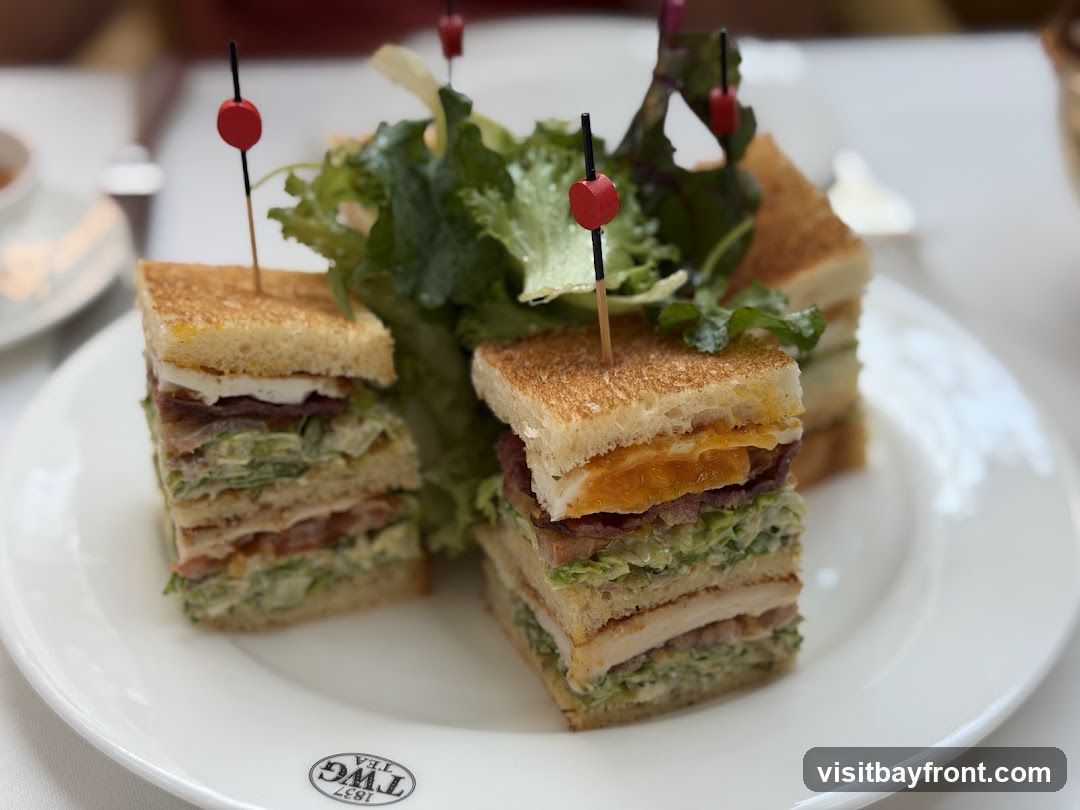 Two club sandwiches are presented on a white plate with a side of salad. The sandwiches are cut into quarters and held together with toothpicks. The plate has a logo that reads "TWG TEA 1837" printed on it.