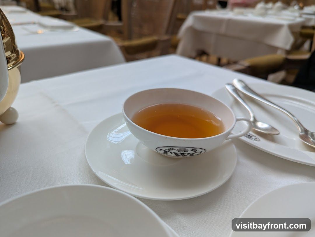 A close-up view of a cup of tea on a saucer, with two spoons resting on a plate to the right. The cup has a logo that appears to be 'TWC'. The setting is formal, with white tablecloths and blurred background suggesting a restaurant or tea room.