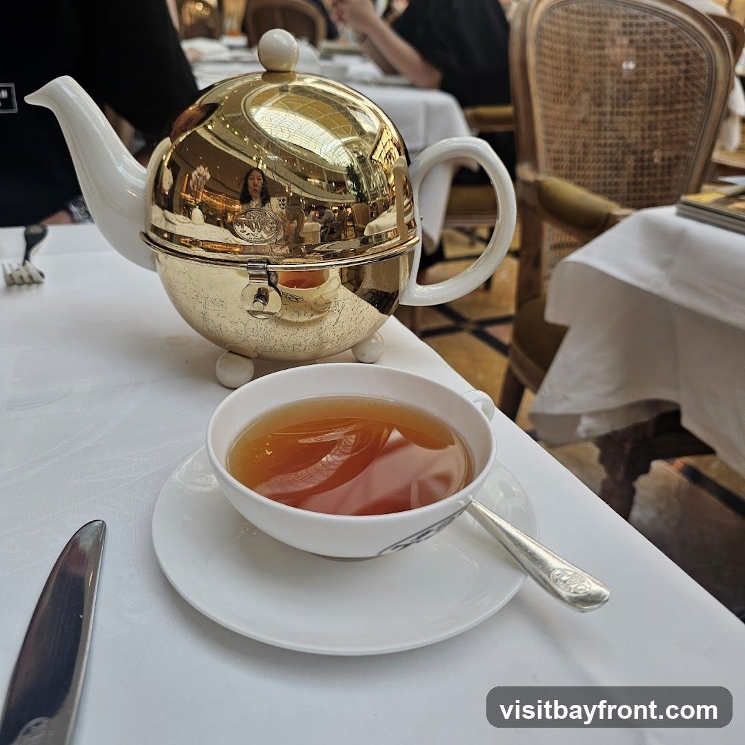 A golden teapot and a cup of tea are set on a white tablecloth. The teapot has a white spout and handle, and its golden body reflects the interior of a room with a domed ceiling and chandeliers. A person is visible in the reflection. A silver knife and spoon are also on the table.