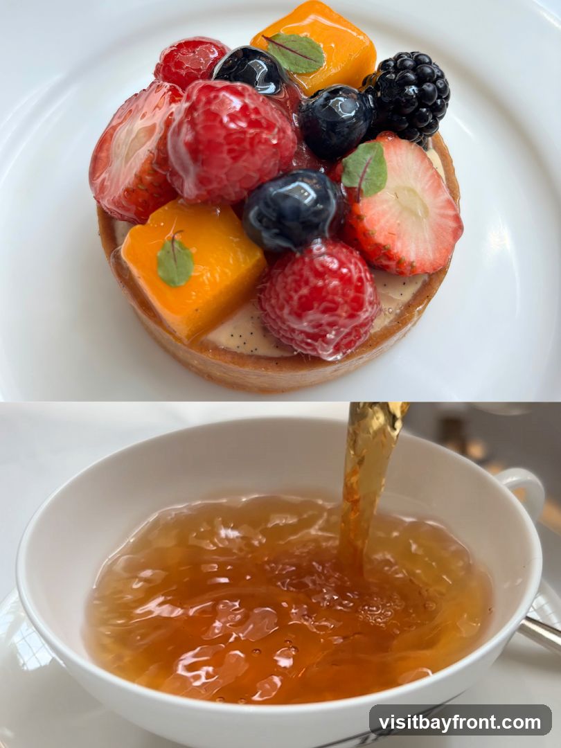 A close-up of a fruit tart topped with strawberries, raspberries, blueberries, blackberries, and mango cubes, served on a white plate. Below, tea is being poured into a white teacup.