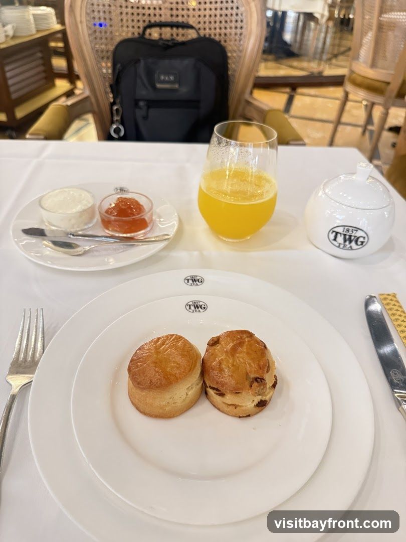 A table setting for afternoon tea, featuring two scones on a plate, a glass of orange juice, a small dish of jam and cream, and a sugar bowl. A black backpack is visible on a chair in the background. The tableware is branded with "TWG TEA 1837".