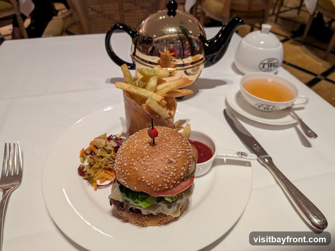 A close-up shot of a meal served on a white tablecloth. In the foreground, a sesame seed bun burger is presented with a side of coleslaw and a small dish of ketchup. Next to it, a cone of french fries is piled high. In the background, a shiny, golden teapot sits beside a white sugar bowl and a cup of tea, all bearing the TWG logo. A knife and fork are also visible on the table.
