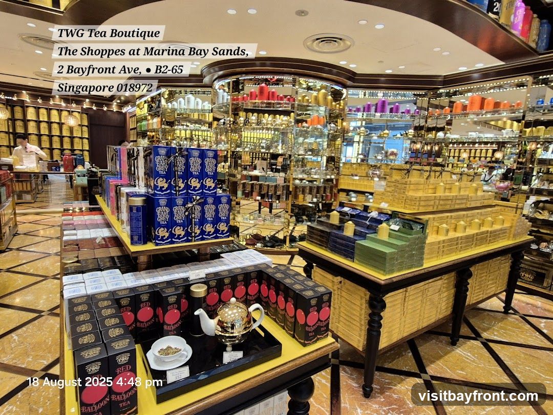 The interior of a TWG Tea Boutique, showcasing a wide selection of teas in decorative packaging. Shelves are filled with numerous tea tins and boxes, with a prominent display of tiered boxes in the center. A tea set with a teapot and cup is visible on a table in the foreground. The store appears well-lit and opulent.