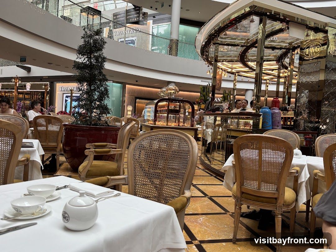 A view of an elegant tea room with white tablecloths, ornate chairs, and a display counter. In the background, a TWG Tea counter is visible with various tea canisters. Other shops, including Tiffany & Co., are seen in the distance within a shopping mall.