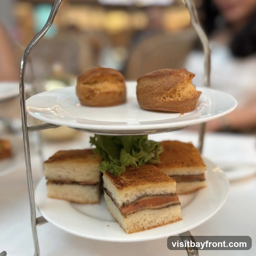 A tiered stand holds an assortment of small sandwiches and scones, presented for afternoon tea. The top tier features two golden-brown scones. The lower tier displays four finger sandwiches, each cut into neat squares, with a garnish of green lettuce. The sandwiches appear to have a filling of thinly sliced salmon or similar. The setting suggests a formal or elegant tea service.