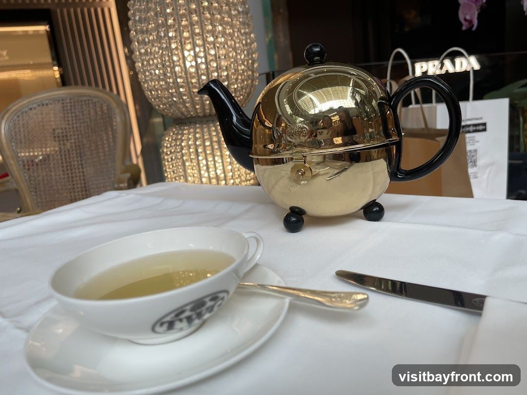 A golden teapot and a cup of tea are set on a white tablecloth. In the background, a Prada shopping bag is visible, along with a decorative lamp and a chair.