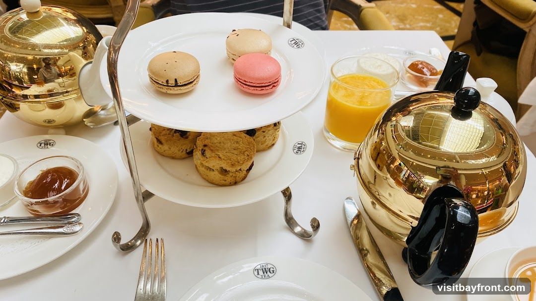 A close-up view of an afternoon tea set, featuring a tiered stand with scones and macarons. A golden teapot, a glass of orange juice, and small bowls of jam and cream are also visible on the white table. The TWG Tea logo is present on the plates.