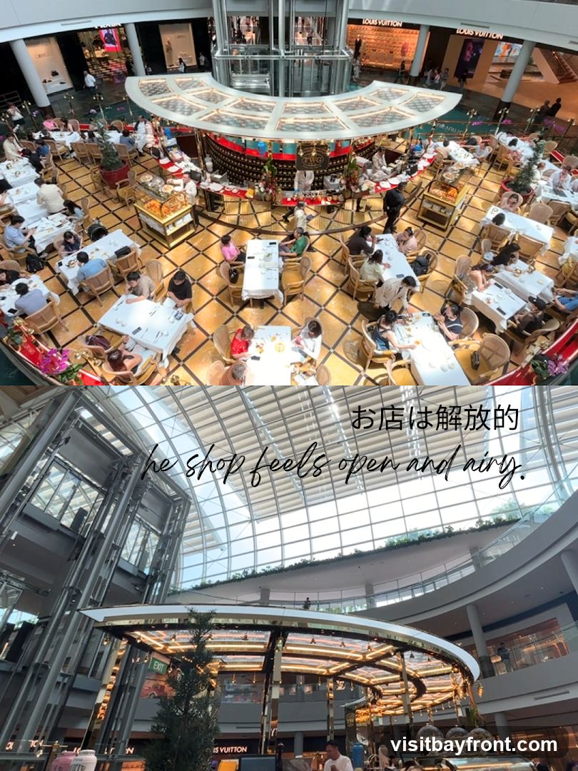 An overhead view of a busy indoor dining area within a large, modern shopping mall. Tables are set with white tablecloths, and patrons are seated, eating and conversing. In the background, high-end retail stores like Louis Vuitton are visible. The lower portion of the image shows a closer view of the mall's architecture, with a glass ceiling and metallic structures. Text overlays read "お店は解放的" and "he shop feels open and airy."