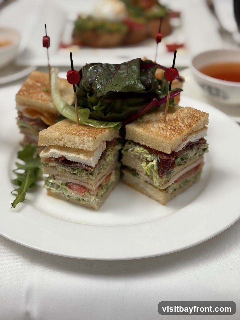 A plate of club sandwiches cut into quarters, garnished with greens and secured with toothpicks. In the background, other plates with food and a cup of tea are visible.