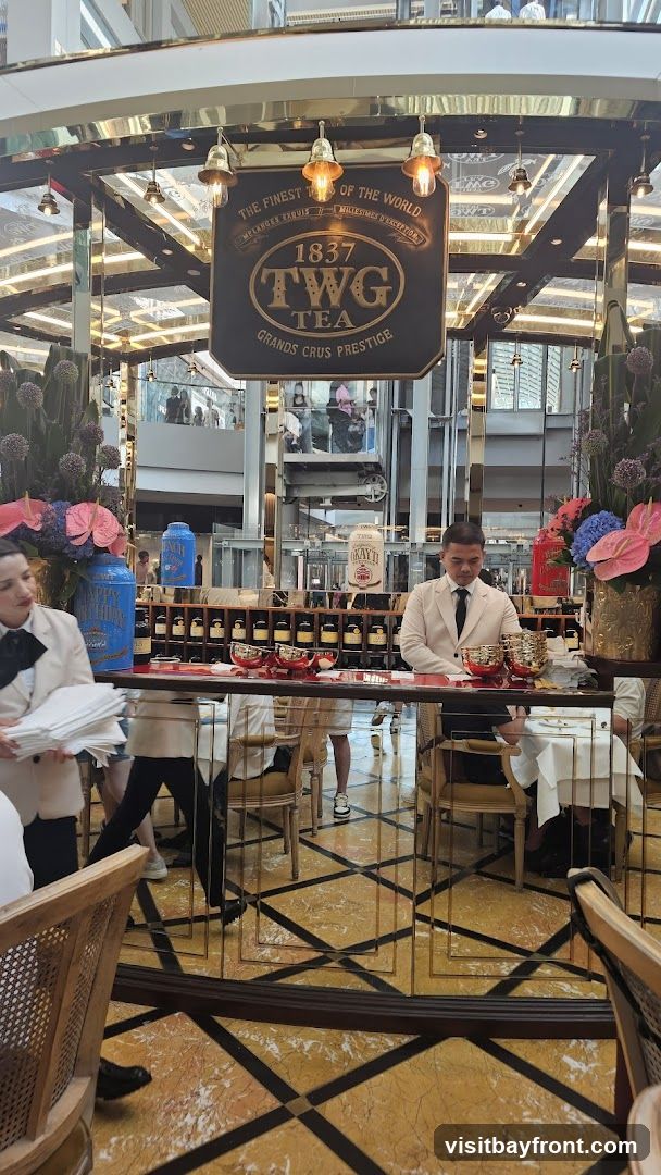 A view inside a TWG Tea establishment, featuring a prominent sign that reads "THE FINEST TEAS OF THE WORLD 1837 TWG TEA GRANDS CRUS PRESTIGE". Staff members in uniform are visible, along with displays of tea canisters and bottles. The interior has a luxurious feel with mirrored surfaces and ornate lighting.