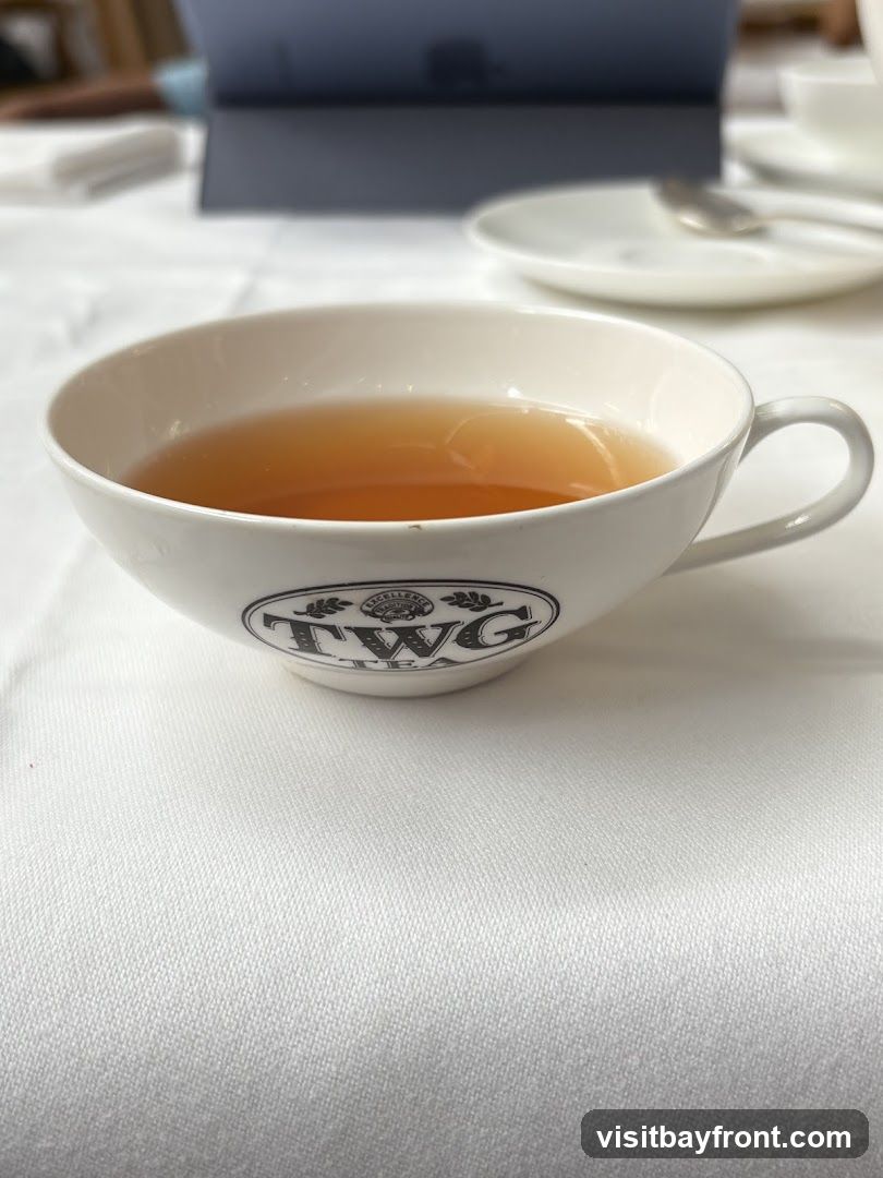 A close-up shot of a white teacup filled with amber-colored tea. The cup has a black logo that reads "TWG TEA" with decorative elements. The cup is placed on a white tablecloth, with a blurred background showing a plate, spoon, and a laptop.