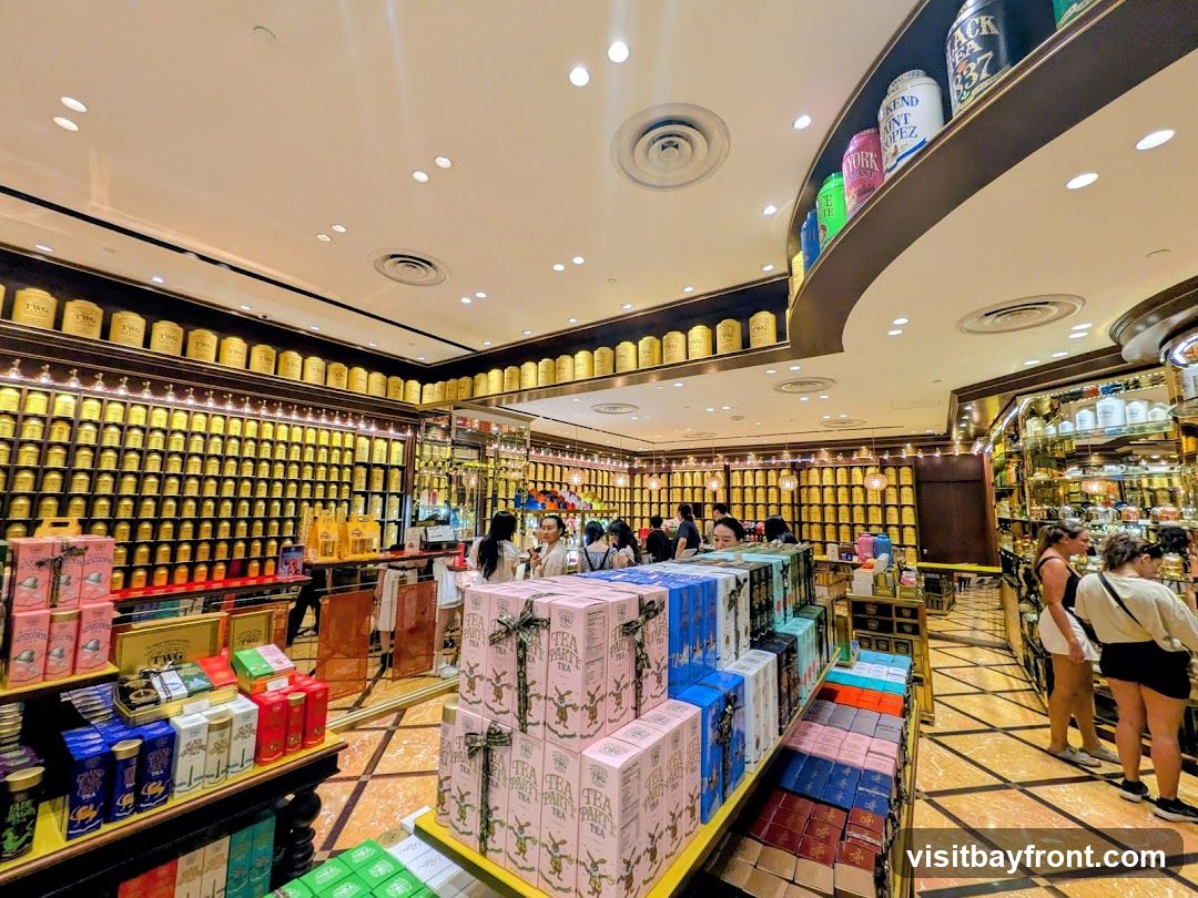 The interior of a tea shop with walls lined with shelves filled with numerous golden tea canisters. In the foreground, there are displays of boxed teas, some with intricate designs. Several customers are browsing the merchandise.