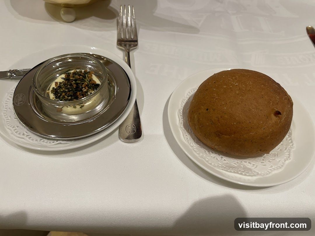 A close-up shot of a bread roll and a small dish of butter with herbs and spices, served on a white tablecloth. A fork is visible to the right of the butter dish. The butter dish has a silver base with the TWG Tea logo embossed on it.