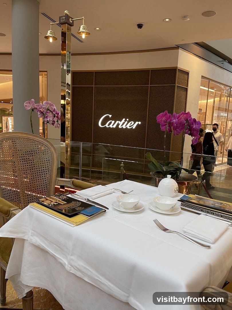 A table is set for tea with white tablecloths, teacups, saucers, and silverware. Two floral arrangements with pink orchids are on display. In the background, the illuminated Cartier logo is visible on a dark textured wall, suggesting a cafe or lounge area within a Cartier store.