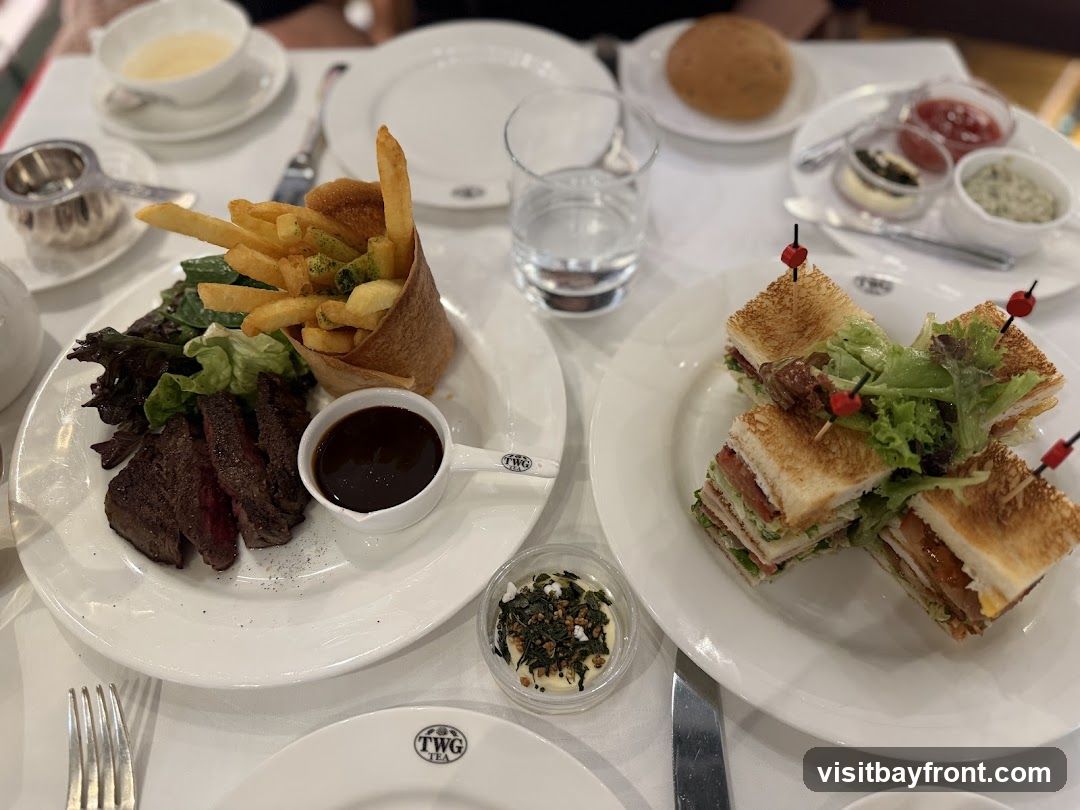A table setting with two plates of food. One plate has sliced steak, fries, salad, and a small dish of sauce. The other plate has several club sandwiches cut in half and garnished with lettuce. There are also small dishes of condiments and a glass of water on the table. The TWG Tea logo is visible on a plate in the foreground.