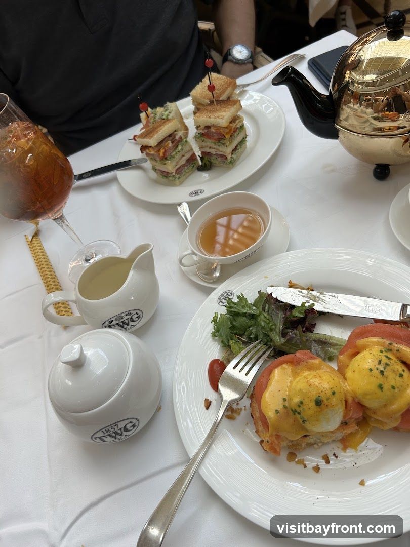 A table set for afternoon tea with various dishes and beverages. On the table are two plates of sandwiches, one with stacked club sandwiches and another with eggs benedict topped with salmon. A teapot, a creamer, a sugar bowl, and a glass of iced tea are also visible. The tableware is branded with "TWG TEA 1837".