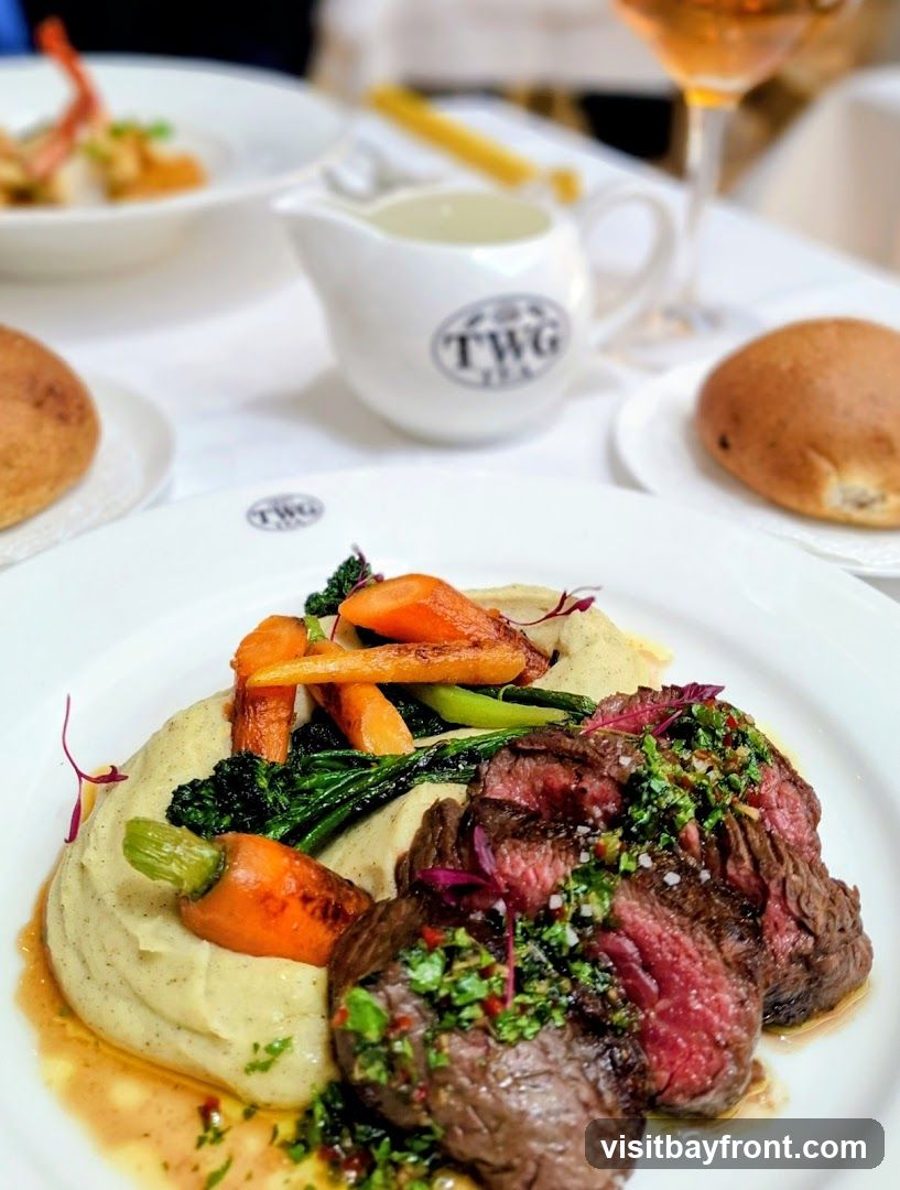 A close-up shot of a steak dish served with mashed potatoes, roasted carrots, and greens. A small pitcher with the TWG Tea logo is visible in the background, along with a glass of amber liquid and a bread roll.