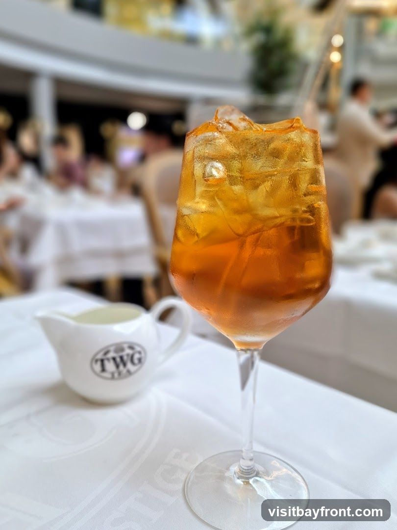 A tall glass of iced tea with ice cubes sits on a white tablecloth. Next to it is a small white creamer with the TWG Tea logo. The background is blurred, showing a restaurant interior with tables and people.