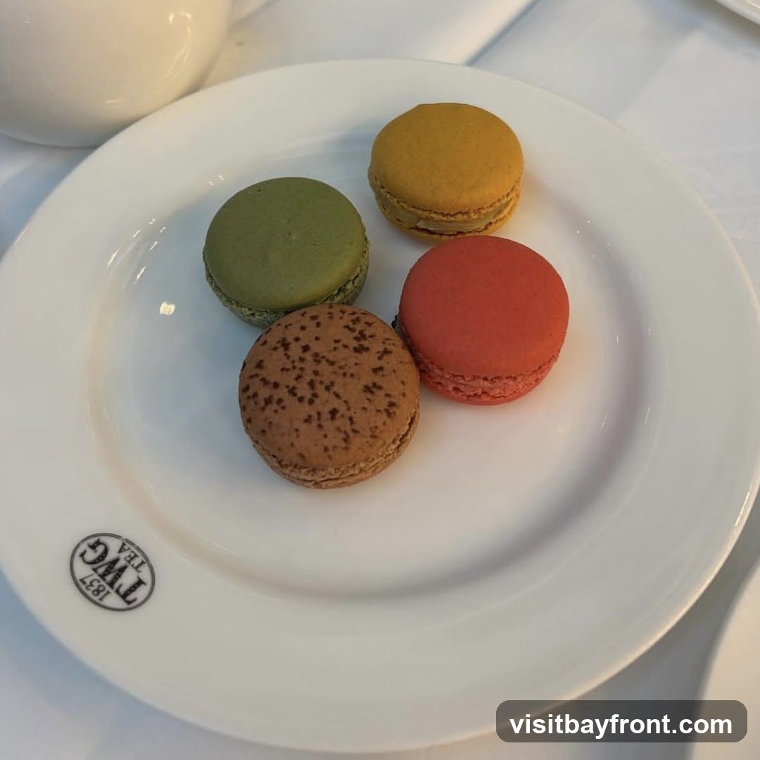 A white plate holds four colorful macarons: green, yellow, red, and brown with speckles. The plate has a logo that reads "TWG TEA 1837" in a circular emblem. A white teapot is partially visible in the upper left corner.