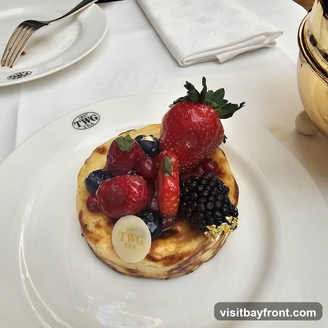 A close-up shot of a berry tart served on a white plate. The tart is topped with fresh strawberries, raspberries, blueberries, and blackberries. A small white chocolate disc with the TWG Tea logo is placed on the tart. The plate has the TWG Tea logo embossed on it. A fork and a folded napkin are visible on the side. The setting appears to be a restaurant or tea house with a white tablecloth.