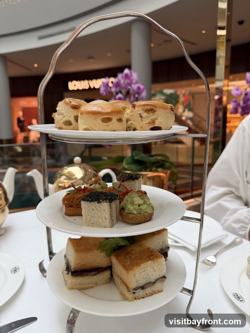 A three-tiered stand filled with various pastries and sandwiches, including scones, mini sandwiches, and tarts. A teapot and teacups are visible on the table. In the background, a Louis Vuitton store is visible with purple orchids.