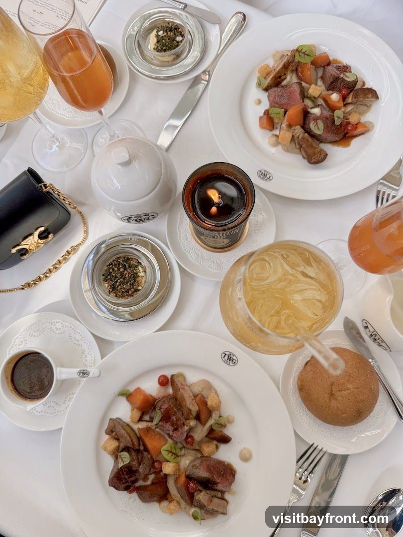 A beautifully set table with multiple plates of food, drinks, and a candle. The dishes appear to be meat with vegetables and sauce. There are also glasses of beverages, a small cup of coffee, and a bread roll. The tableware is branded with "TWG TEA 1837". A black handbag with a gold chain is also visible on the table.