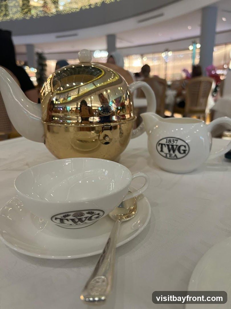 A close-up shot of a white teacup and saucer with a silver spoon, a white milk jug, and a golden teapot. The TWG Tea logo is visible on the teacup and milk jug. The background shows a blurred view of a shopping mall with people dining.
