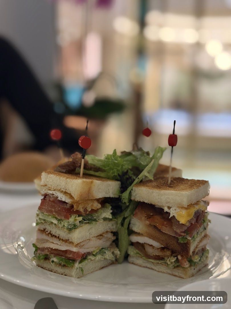 A tall club sandwich cut in half and served on a white plate. The sandwich is made with toasted bread, layers of turkey, bacon, lettuce, and tomato, and is held together with toothpicks. The background is blurred and appears to be an indoor setting with soft lighting.