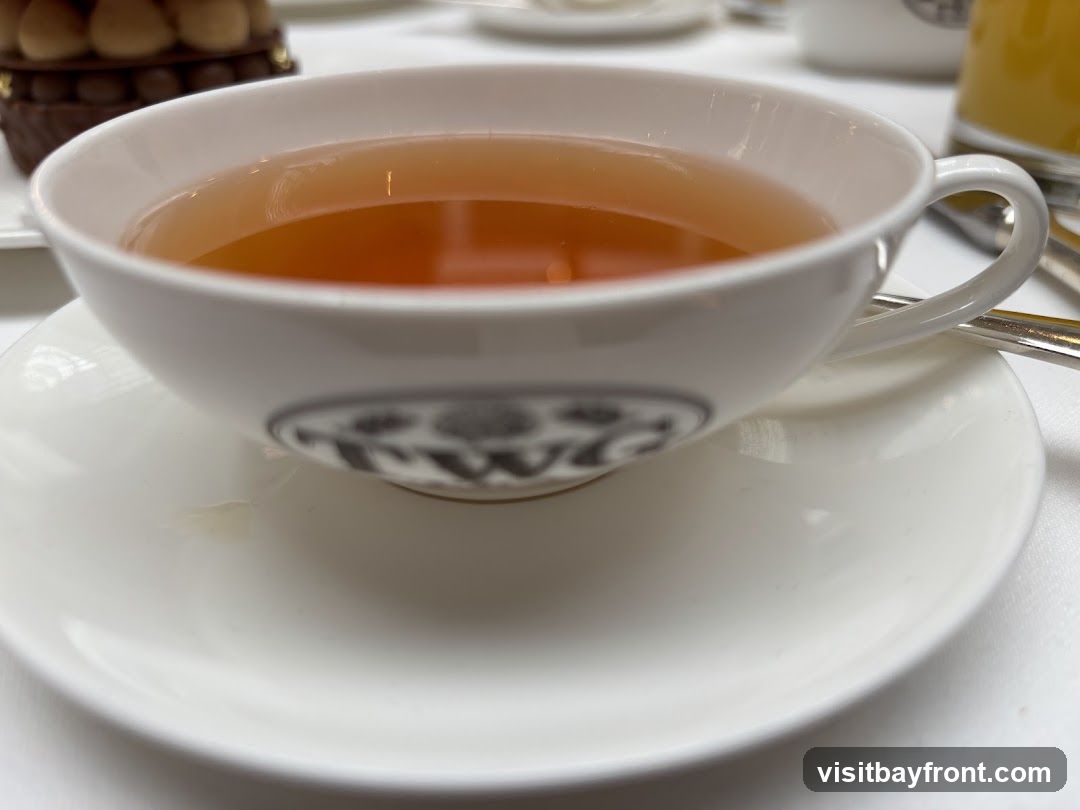 A close-up shot of a white teacup filled with amber-colored tea, resting on a matching saucer. The cup has a dark logo visible on its side. In the background, a dessert and a glass of orange juice are out of focus.