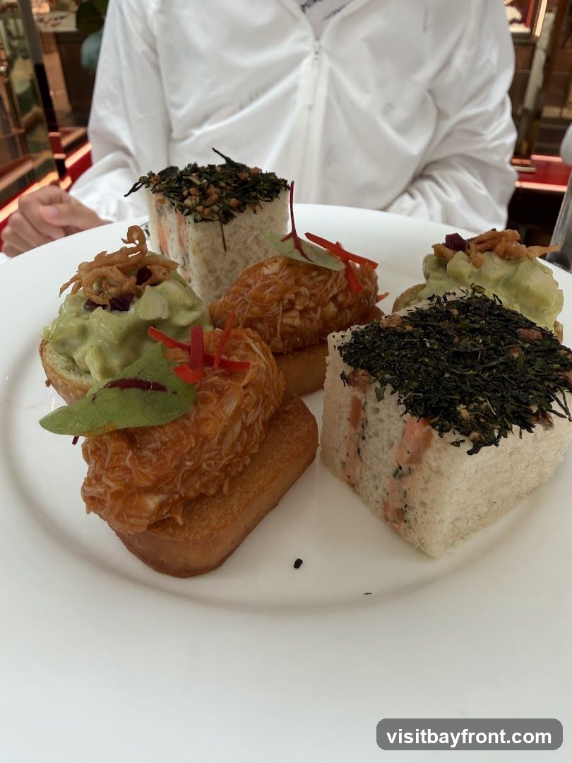 A white plate holds an assortment of small, savory bites. There are square sandwiches topped with dark green herbs and what appears to be crispy fried onions. Other items include small toasts topped with a green spread, possibly avocado, and garnished with red chili and crispy fried onions. There are also pieces of what looks like shredded crab meat on small toasts, decorated with red chili and a green leaf. A person wearing a white jacket is visible in the background.
