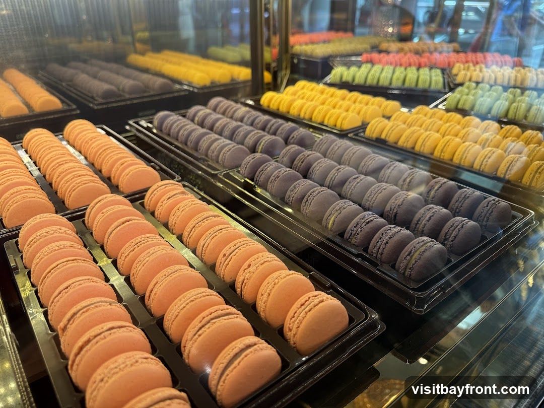 A display case filled with rows of colorful macarons. The macarons are arranged in plastic trays, with visible colors including peach, purple, yellow, green, and pink. The display appears to be in a bakery or confectionery shop.