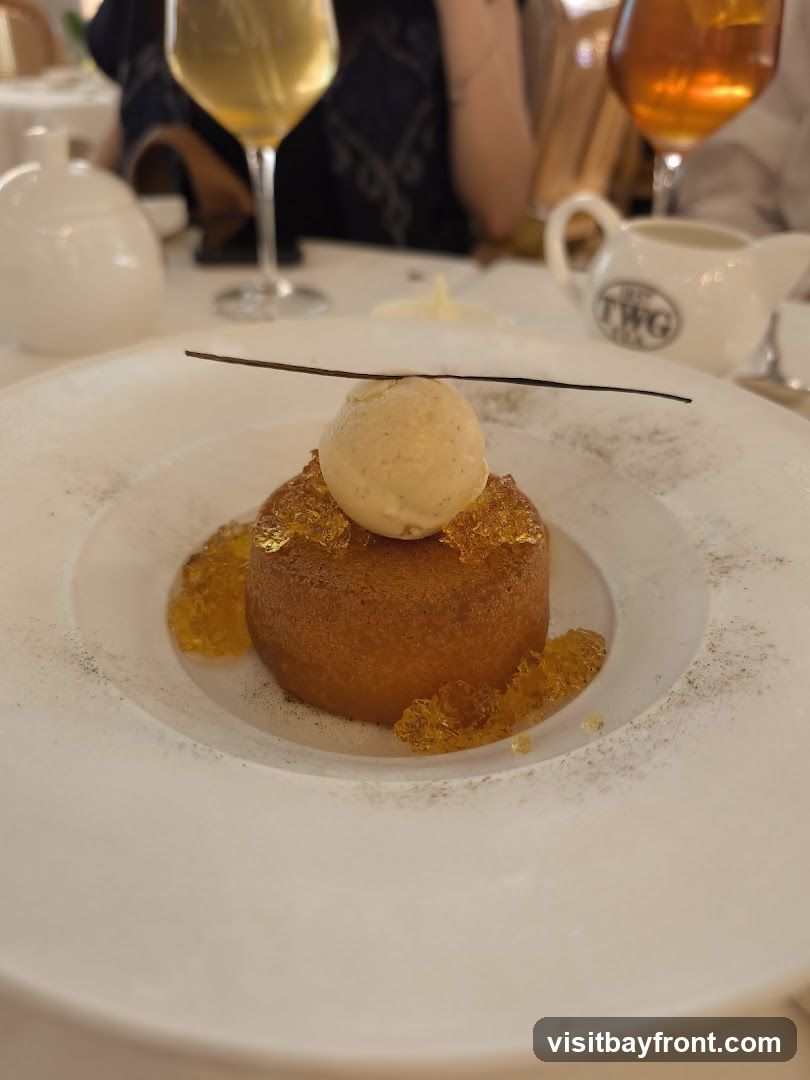 A close-up shot of a dessert on a white plate. The dessert is a small, round cake topped with a scoop of vanilla ice cream and a thin dark stick. It is surrounded by amber-colored jelly pieces and dusted with a dark powder. In the background, there are glasses of drinks and a white creamer with the TWG Tea logo.