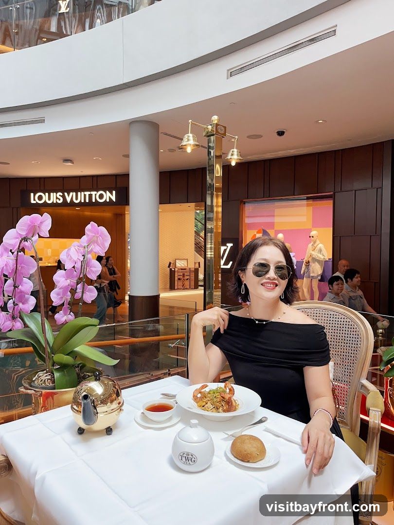 A woman sits at a table set with a white tablecloth, enjoying a meal. In front of her is a plate of pasta with shrimp, a cup of tea, a sugar bowl, and a bread roll. A golden teapot sits to her left. Behind her, the Louis Vuitton store is visible, suggesting the restaurant is located within a shopping mall. The ambiance appears elegant and sophisticated.