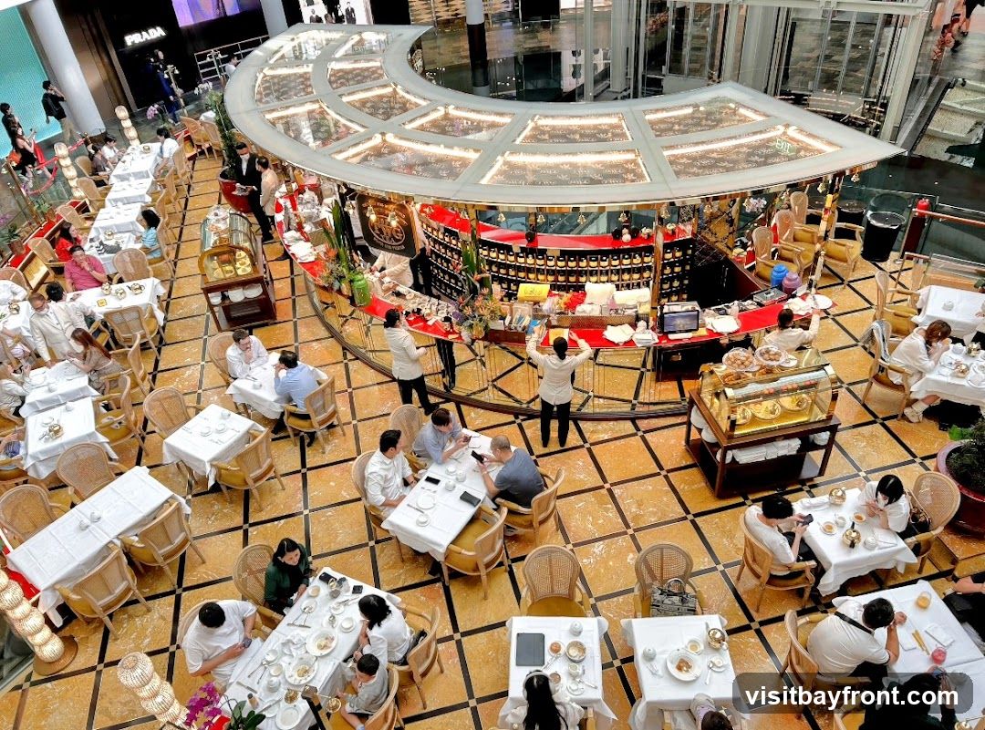 An overhead view of a bustling tea room or cafe, featuring a circular counter with staff serving patrons. Numerous tables are set with white tablecloths and chairs, occupied by diners enjoying their meals and drinks. The decor is elegant, with ornate lighting and a patterned floor. In the background, a Prada store is visible, suggesting the location is within a high-end shopping mall.